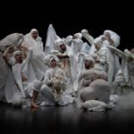 Gruppo di danzatori vestiti di bianco e ricoperti di teli e plastiche nella pi